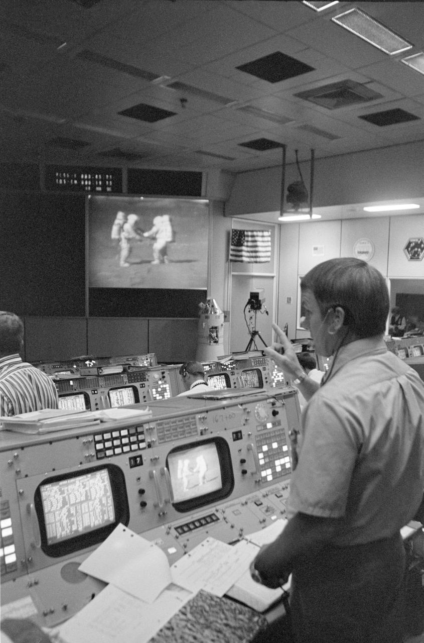 Control room with personnel monitoring a screen displaying astronauts, vintage setting.
