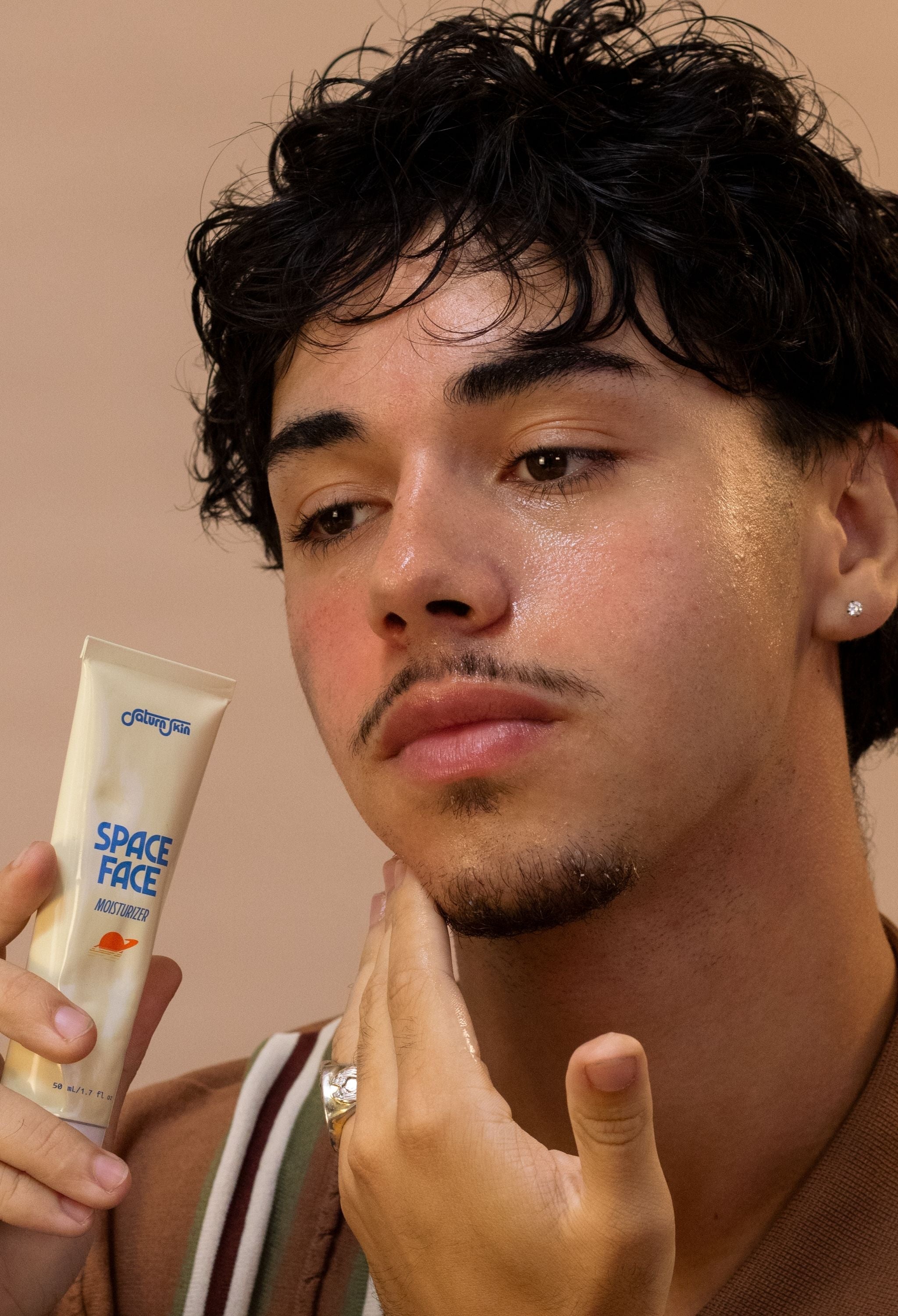 Man with dark curly hair and mustache holding a tube of Space Face Moisturizer by Saturn Skin and applying to his face against a beige background